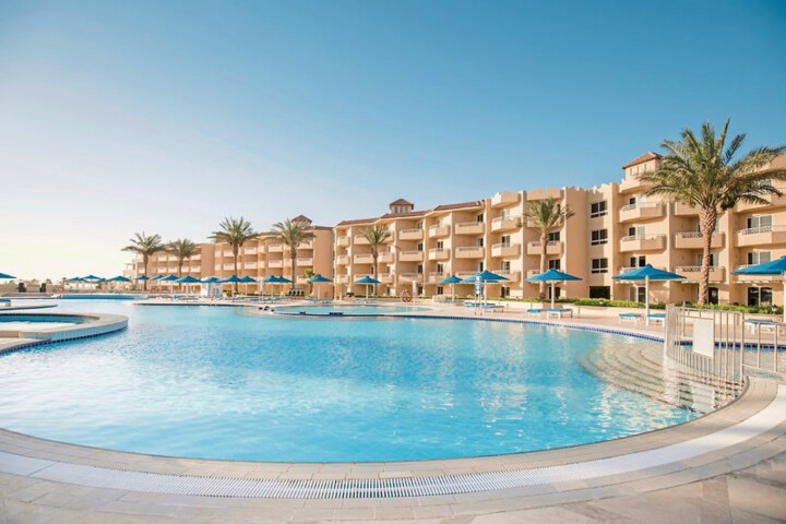 Großer, geschwungener Pool mit blauen Sonnenschirmen und Palmen vor einem mehrstöckigen Hotelgebäude unter klarem Himmel.