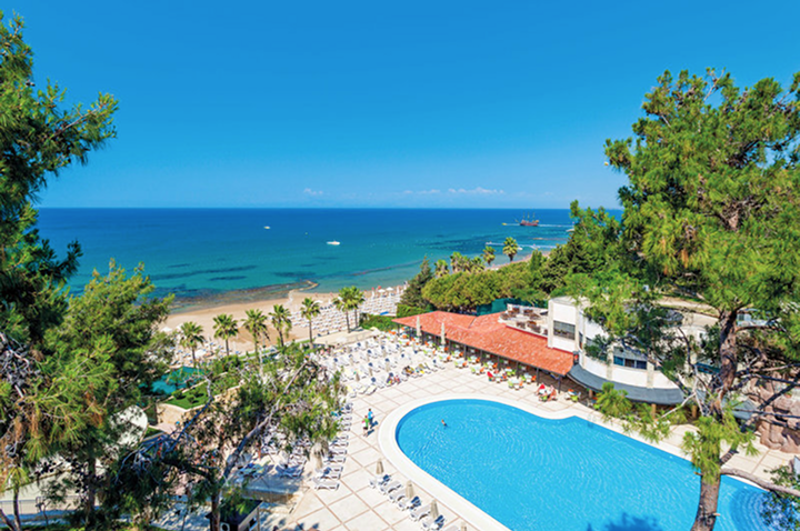 Blick von oben auf einen Pool mit Liegestühlen und Sonnenschirmen, umgeben von Bäumen, mit Strand und Meer im Hintergrund.