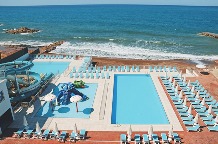 Ein großer Pool und ein Strand mit Liegen und Sonnenschirmen, umgeben von Palmen und strahlend blauem Himmel.