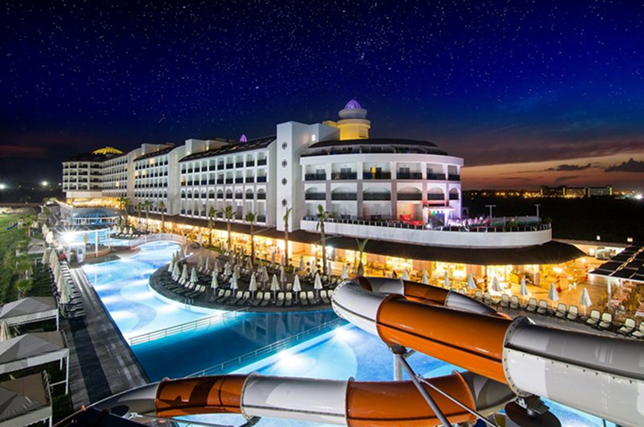 Großes Hotelgebäude mit beleuchtetem Pool und Wasserrutsche bei Nacht unter klarem Sternenhimmel.
