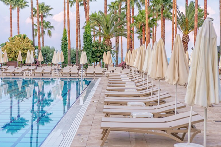 Reihe von Liegestühlen mit geschlossenen Sonnenschirmen neben einem Schwimmbecken und Palmen im Hintergrund.
