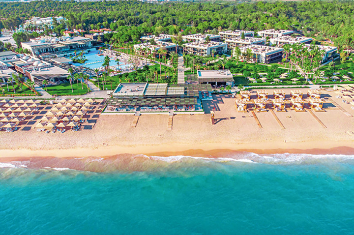Aerialansicht eines Resorts und Strandes, das Hotel ist sichtbar, umgeben von türkisfarbenem Wasser und grünen Bäumen.