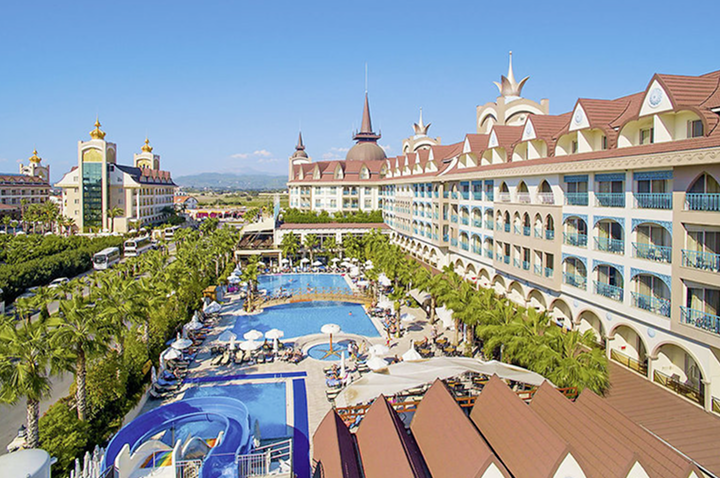 Ein großes Hotel mit einem einladenden Swimmingpool, umgeben von Palmen und einer sonnigen Terrasse.