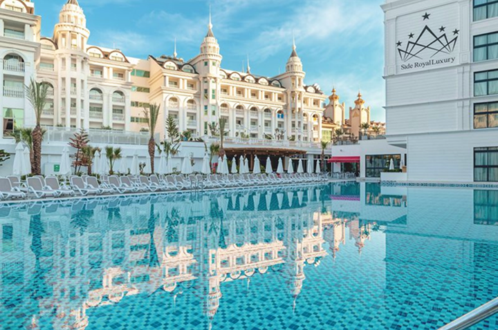 Großer Außenpool mit türkisblauem Wasser vor einem weißen, prunkvollen Hotelgebäude im klassischen Stil mit Türmchen und Balkonen.