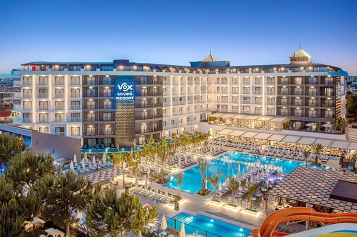 Großes mehrstöckiges Hotel mit beleuchtetem Schriftzug und mehreren Pools mit Liegen und Sonnenschirmen davor.