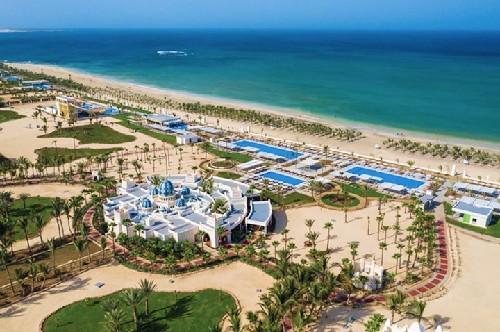 Luftaufnahme eines Resorts mit weiß-blauen Gebäuden, mehreren Pools, Palmen und einem Sandstrand am Meer