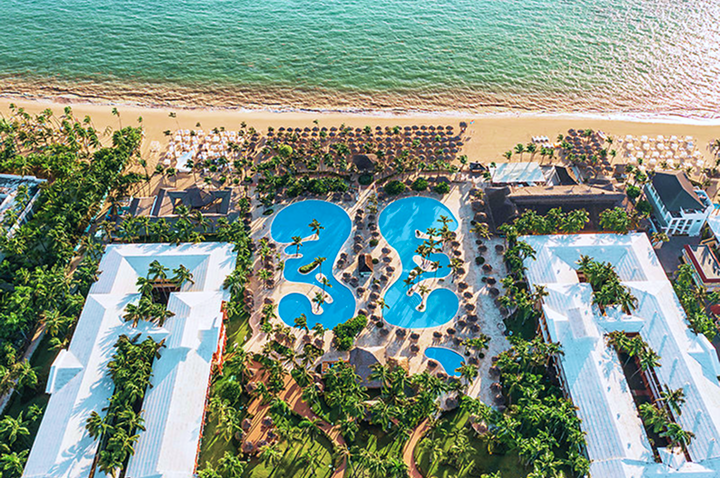 Luftaufnahme eines Resorts mit zwei großen, geschwungenen Pools, umgeben von Palmen und weißen Gebäuden am Strand mit türkisfarbenem Meer. Luftaufnahme eines Resorts mit zwei großen, geschwungenen Pools, umgeben von Palmen und weißen Gebäuden am Strand mit türkisfarbenem Meer.