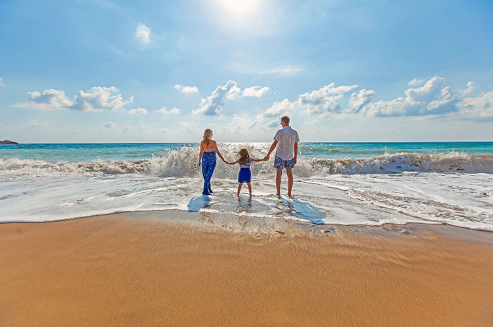 Familie mit zwei Erwachsenen und einem Kind hält sich am Strand an den Händen und blickt aufs Meer mit Wellen und bewölktem Himmel.