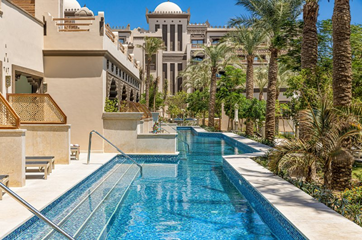 Langer Pool mit Treppen und Handläufen neben modernen Gebäuden und Palmen unter blauem Himmel