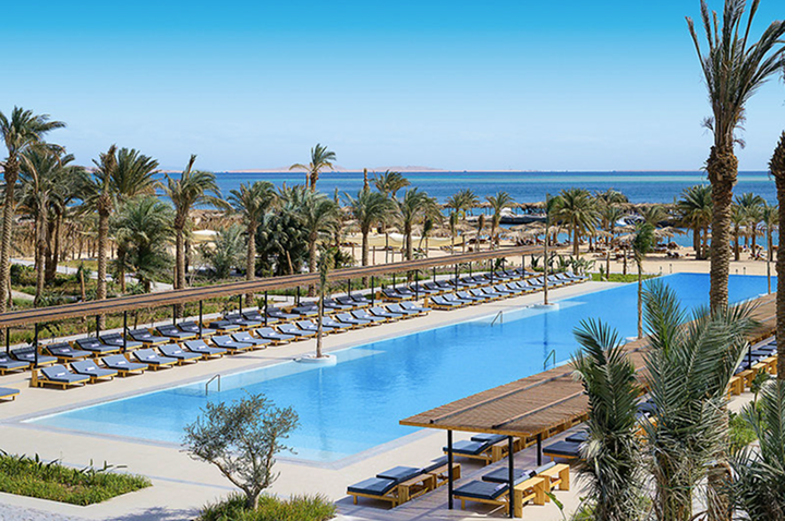 Großer rechteckiger Pool mit Liegen und Palmen in einer Strandhotelanlage am Meer.