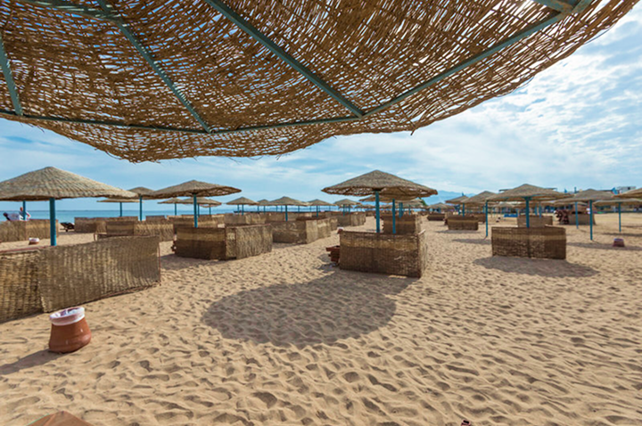 Strand mit mehreren Strohhut-Sonnenschirmen und windgeschützten Sitzbereichen aus geflochtenem Material auf Sand unter blauem Himmel mit Wolken
