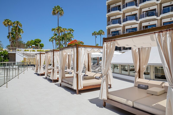 Reihe von überdachten Liegebetten mit Vorhängen auf einer Hotelterrasse neben einem mehrstöckigen Gebäude und Palmen im Hintergrund.