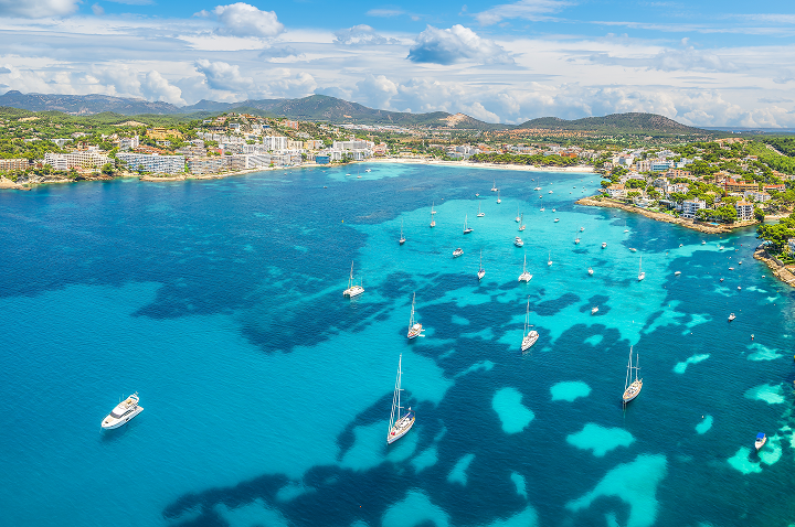 Luftaufnahme einer Bucht mit klarem Wasser, mehreren Segel- und Motorbooten sowie einer Küstenstadt im Hintergrund.