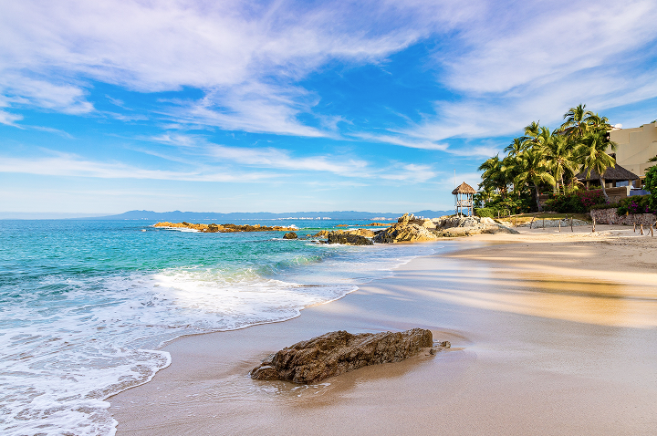 Sandstrand mit Felsen im Vordergrund, Palmen und einem kleinen Aussichtsturm am Rand, ruhiges Meer und bewölkter Himmel.
