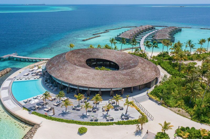 Rundes, strohgedecktes Gebäude mit Innenhof an einem Strand mit Palmen und über Wasser gebauten Bungalows.