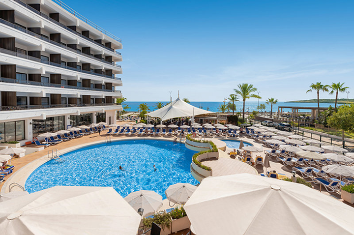 Hotelpool mit Liegestühlen und Sonnenschirmen vor mehrstöckigem Hotelgebäude und Meer im Hintergrund.