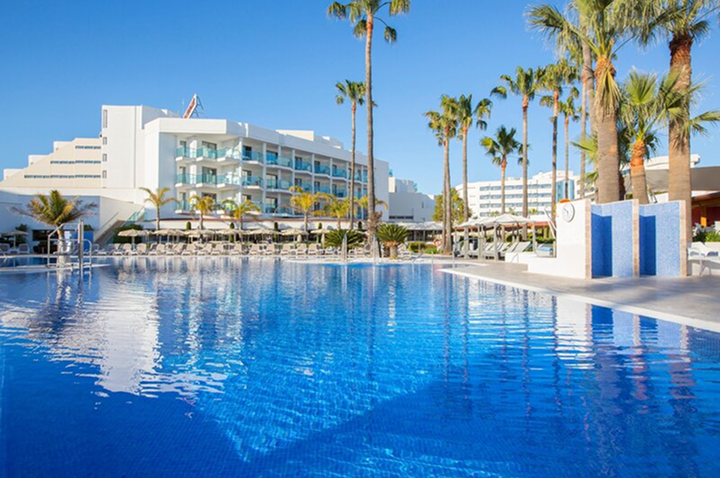 Ein großer blauer Swimmingpool vor einem Hotel, umgeben von Liegen und Sonnenschirmen unter klarem Himmel.