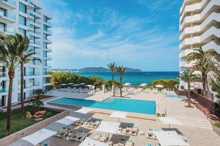 Blick auf einen rechteckigen Pool mit Liegen und Sonnenschirmen zwischen zwei mehrstöckigen Hotels, im Hintergrund das Meer und eine Insel.