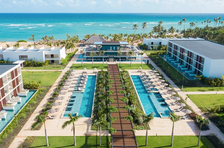 Blick auf ein Resort mit zwei langen Pools, Liegestühlen, Palmen und dem Meer im Hintergrund.