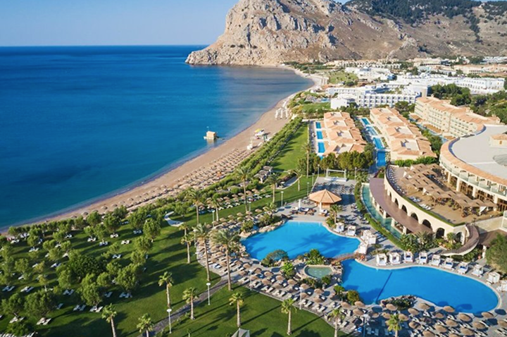 Luftaufnahme eines Resorts mit mehreren Pools, Liegestühlen, Grünflächen und einem Strand an einer felsigen Küste.