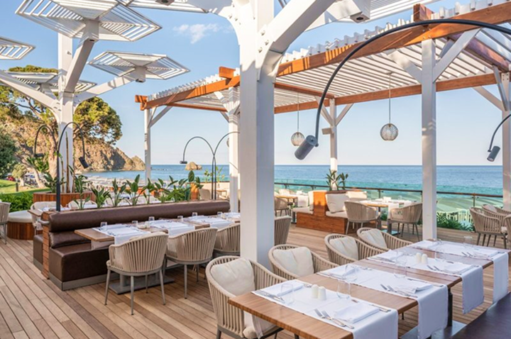 Offenes Restaurant mit Holzboden, weißen Tischen und Stühlen unter weißer Pergola mit Blick auf das Meer und eine Felsklippe.