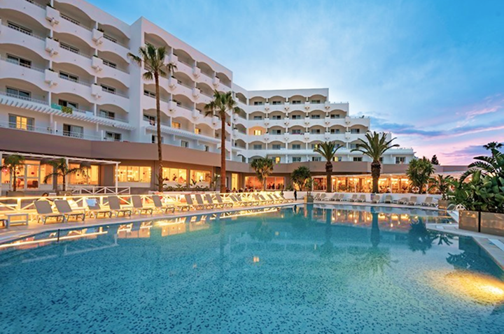 Großes Hotelgebäude mit mehreren Balkonen, beleuchteter Pool und Liegestühle am Abend unter blauem Himmel mit Wolken.