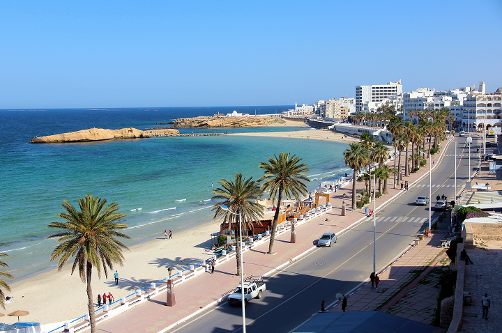 Küstenpromenade mit Palmen, Strand und mehreren Gebäuden am Meer unter klarem Himmel.